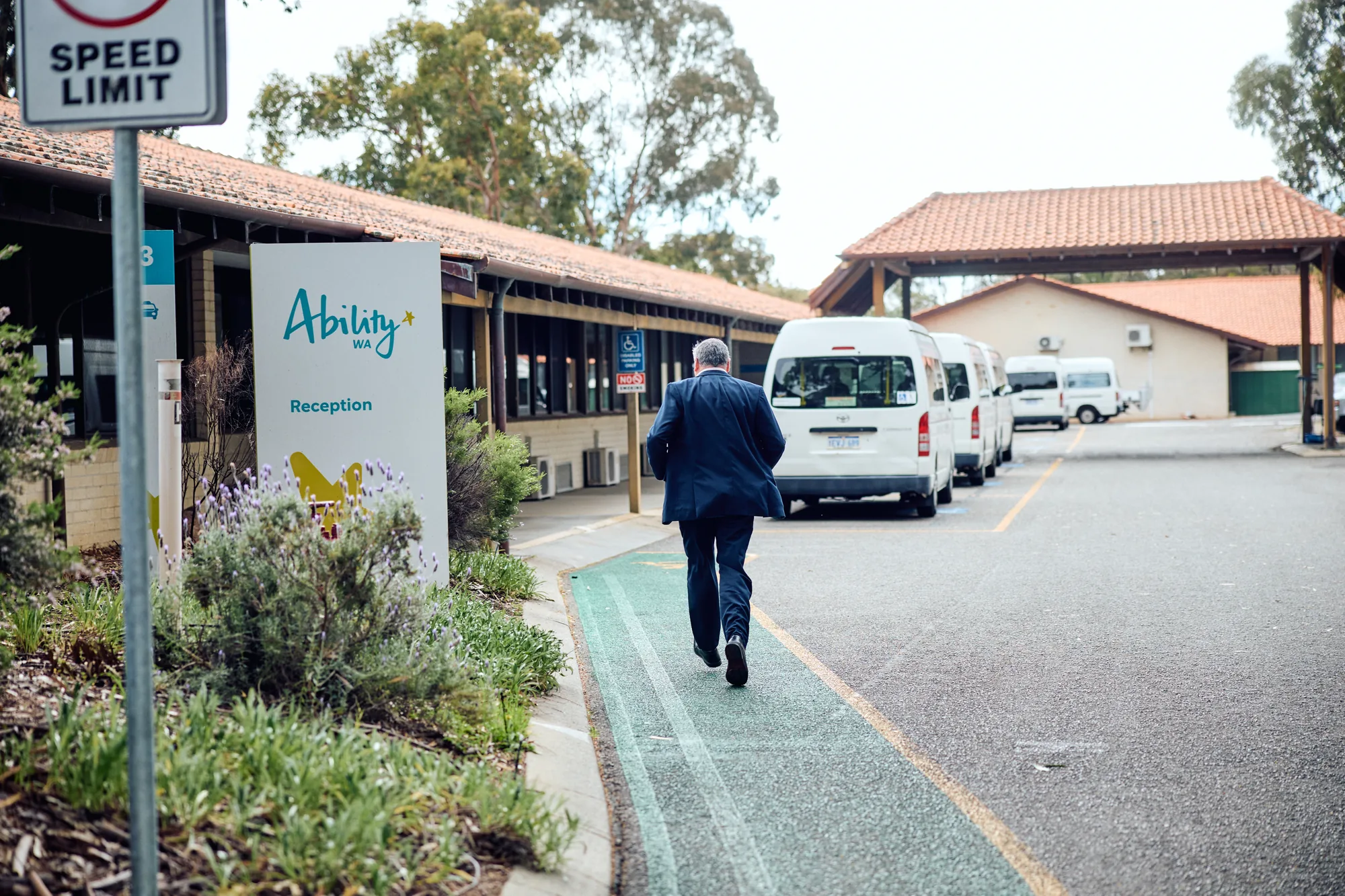 A man in a suit walking up the path to the Ability WA reception entrance, past a row of parked white minibuses