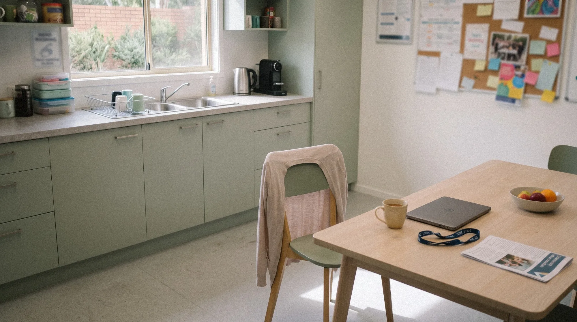An empty staff room with sage green kitchen cabinets, a cardigan over a chair, and a mug of tea, laptop and lanyard on a timber table